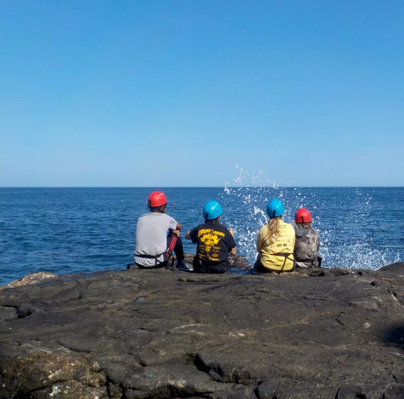 Four people wearing helmets are sitting on a rocky outcrop, looking out at the ocean. The sky is clear and blue, and the water is a deep blue color. There are some waves crashing against the rocks in the distance. The people are all wearing different colored helmets, and they appear to be enjoying the view.